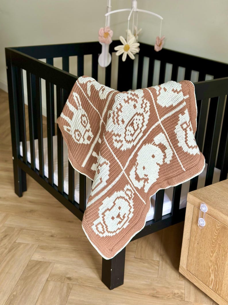 K&ouml;nnte beinhalten: Eine handgefertigte Babydecke aus H&auml;kelarbeit in Braun und Wei&szlig; mit Tiermustern. Die Decke liegt &uuml;ber der Seite eines schwarzen Kinderbetts. &Uuml;ber dem Bett h&auml;ngt ein Mobile mit Blumen- und Sternformen.