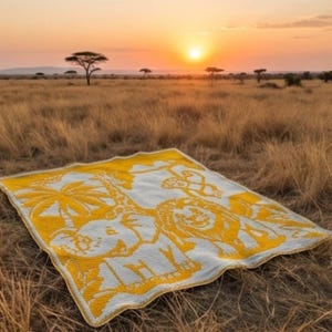 May include: A white and yellow blanket with an African safari animal design is spread on the ground. The blanket features a giraffe, lion, and elephant. The background shows a sunset over a grassy plain with trees.