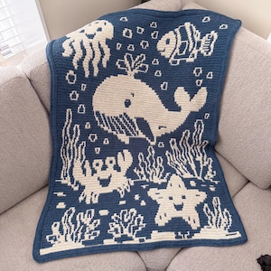 May include: A blue and white crocheted blanket featuring an underwater scene. The design includes a whale, fish, jellyfish, crab, and starfish. The blanket is folded over a gray sofa.