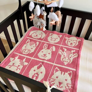 Può includere: Una coperta per neonati all'uncinetto rosa e bianca con facce di animali, tra cui un cane, un maiale, una pecora, una zebra, un coniglio, una giraffa, un cavallo, una mucca e un gatto. La coperta è in una culla marrone scuro, con una giostrina appesa sopra.