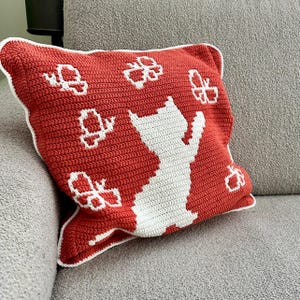 May include: A red crochet pillow with a white cat silhouette and white butterfly accents. The pillow has a white border and is resting on a gray couch. This handmade pillow is a decorative home accent.