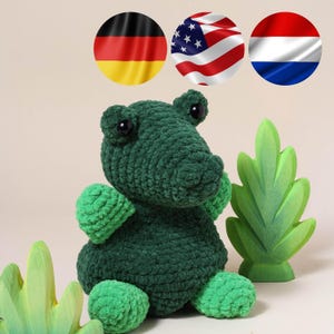 May include: A handmade, dark green crochet alligator plush toy with lighter green accents on its feet and arms. The alligator has black button eyes and is sitting next to a green leaf decoration. The image also includes the flags of Germany, the United States, and the Netherlands.