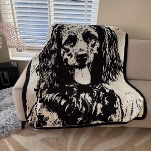 May include: A rectangular crochet blanket featuring a high-contrast black and white graphic portrait of a long-haired dog with its tongue hanging out. The design is created with intricate yarn stitches, finished with a solid black border edge.