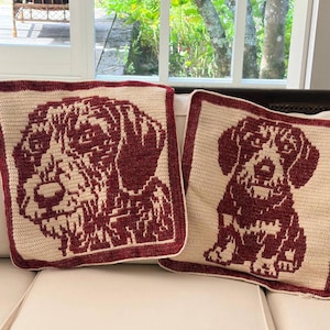 Könnte beinhalten: Zwei quadratische Häkelkissen mit einem Hunde-Design. Die Kissen haben einen cremefarbenen Hintergrund mit einem weinroten Hundeporträt. Das Bild des Hundes ist von einem weinroten Rand umgeben. Die Kissen liegen auf einem weißen Sofa.