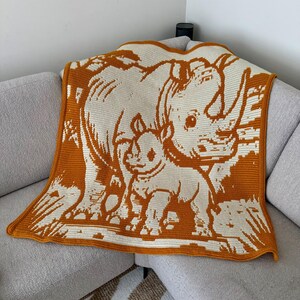 May include: A crocheted blanket featuring a rhino and calf in shades of cream and orange. The blanket is draped over a gray sofa. The design depicts the rhinos in a stylized, graphic manner, suitable for a nursery or child's room.