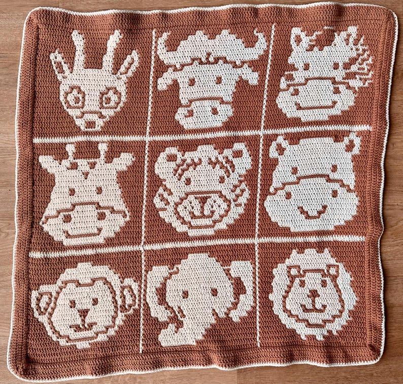 K&ouml;nnte beinhalten: Eine handgefertigte H&auml;keldecke mit einem Raster aus neun Tiergesichtern in Braun und Creme. Zu den Tieren geh&ouml;ren eine Giraffe, ein B&uuml;ffel, ein Tiger, ein Nilpferd, ein L&ouml;we und ein Elefant. Die Decke hat einen wei&szlig;en Rand.