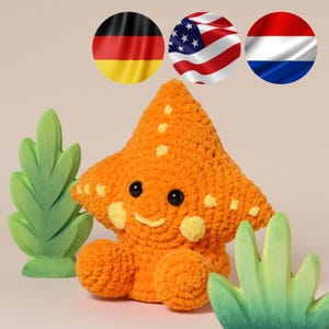 May include: An orange crocheted star-shaped plush toy with yellow spots, black eyes, and a smiling mouth. The toy has two small orange spheres at the bottom. Two green wooden leaf decorations are on either side of the toy.