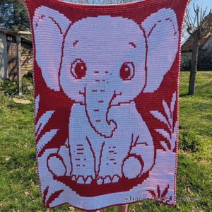 May include: A red and white crocheted blanket featuring a large, smiling elephant. The elephant is white with red accents, including large ears and big eyes. The blanket has a red border and a repeating pattern. The blanket is held up outdoors.