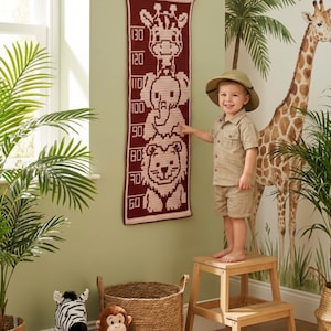 Puede incluir: Un medidor de altura de ganchillo burdeos y crema con animales, que mide hasta 130 cm. Presenta una jirafa, un elefante y un león. Un niño con ropa de safari está de pie sobre un taburete de madera.