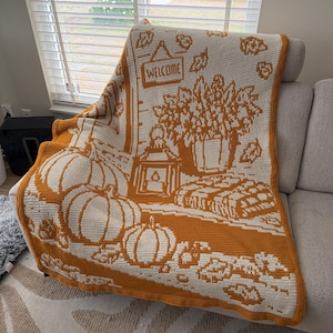 May include: A rectangular crochet blanket featuring a two-tone orange and cream design. The pattern depicts a porch scene with pumpkins, a lantern, a potted plant, and a sign that reads WELCOME, surrounded by scattered autumn leaves.