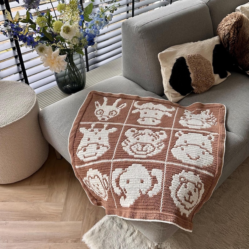 K&ouml;nnte beinhalten: Eine braun-wei&szlig;e H&auml;keldecke mit einem Raster von Tiergesichtern, darunter eine Giraffe, ein Elefant und ein L&ouml;we. Die Decke liegt auf einem grauen Sofa, im Hintergrund eine Vase mit Blumen. Die Decke ist etwa 100 cm gro&szlig;.