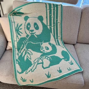 May include: A crocheted blanket featuring a panda bear and cub in teal and white. The blanket has a teal border and depicts bamboo stalks and leaves. The panda and cub are the central focus of the design, with the panda holding bamboo.