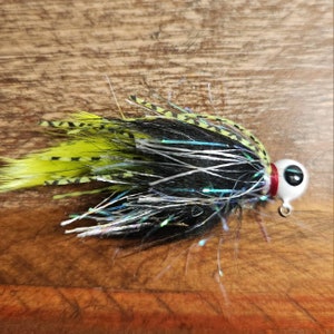 May include: Black and yellow fishing lure with a white and red eye. The lure has a long, flowing tail of black, white, and green fibers.