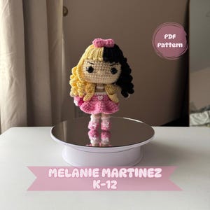 May include: Crocheted doll of Melanie Martinez from her K-12 album. The doll is wearing a pink dress with a heart on the front and a black and white bow in her hair. The doll is standing on a white turntable with a mirrored surface.