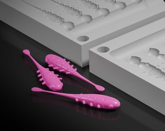May include: A gray plastic mold for making fishing lures. The mold has six cavities, each shaped like a small, segmented fish. The mold is labeled "Slammer Finesse 3.5 cm."  There are also three pink fishing lures made from the mold.