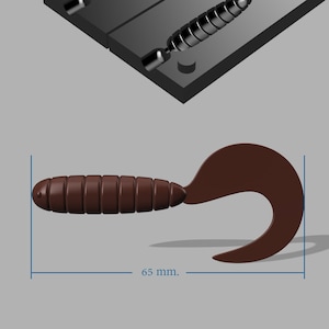 Może przedstawiać: Brązowa przynęta wędkarska z zakrzywionym ogonem, o wymiarach 6,5 cm. Obraz przedstawia również formę do tworzenia przynęty. Przynęta ma żebrowany korpus i zakrzywiony ogon. Forma jest ciemnoszara.