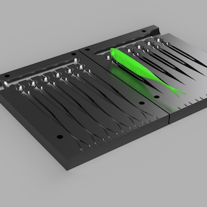 May include: A black, two-part mold for making fishing lures. The mold has multiple cavities in the shape of a fish. A bright green, finished lure rests in one of the cavities. The mold is made of a dark, matte material.