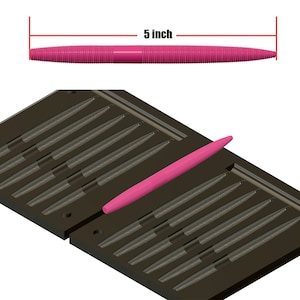 May include: A pink, ribbed fishing lure, 12.7 cm long, is shown on a dark grey mould. The mould has multiple cavities for creating similar lures. The lure is a vibrant pink colour, designed for attracting fish.