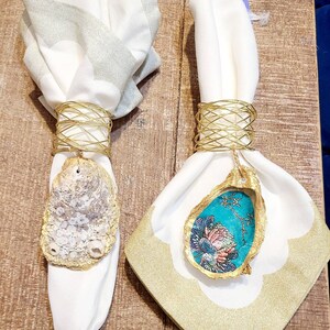May include: Two white napkins with gold wire napkin rings. One napkin ring has a natural seashell attached, the other has a blue and gold seashell with a floral design.