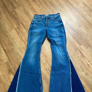 Puede incluir: Un par de pantalones vaqueros de campana azul denim con un panel de tela azul oscuro contrastante en el exterior de cada pierna. Los vaqueros tienen cierre de bot&oacute;n y dobladillo deshilachado.