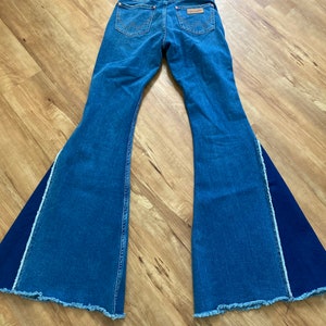 Puede incluir: Un par de pantalones vaqueros de campana azul denim con un panel de tela azul oscuro contrastante en el exterior de cada pierna. Los vaqueros tienen un dobladillo deshilachado y un parche de cuero marr&oacute;n en la parte posterior con el logotipo de Wrangler.