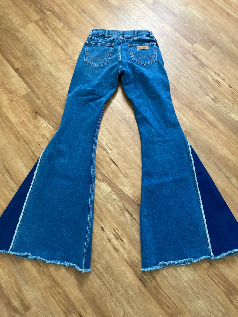 Puede incluir: Un par de pantalones vaqueros de campana azul denim con un panel de tela azul oscuro contrastante en el exterior de cada pierna. Los vaqueros tienen un dobladillo deshilachado y un parche de cuero marr&oacute;n en la parte posterior con el logotipo de Wrangler.