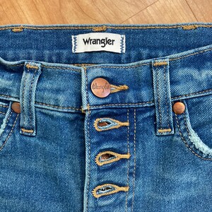 Puede incluir: Vaqueros azules de mezclilla con cierre de bot&oacute;n y una etiqueta de la marca Wrangler. Los vaqueros tienen un aspecto desgastado y un lavado claro.