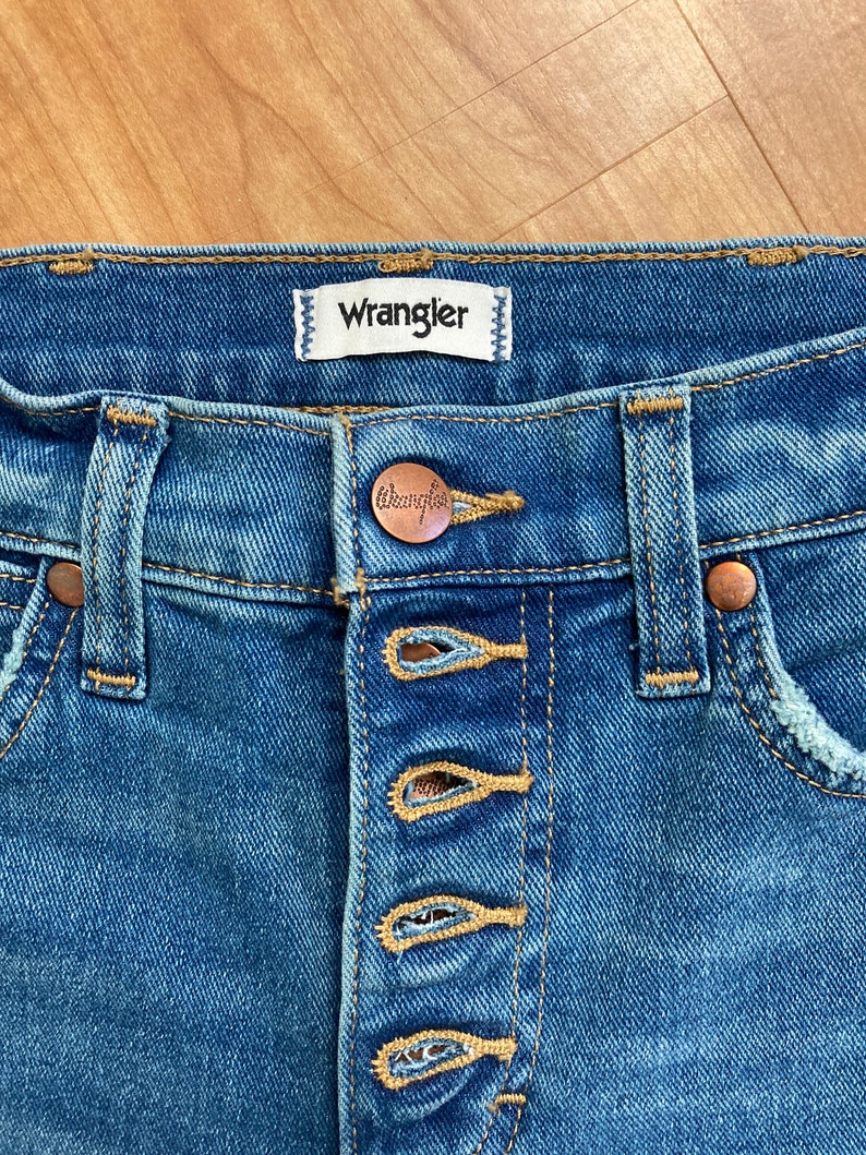 Puede incluir: Vaqueros azules de mezclilla con cierre de bot&oacute;n y una etiqueta de la marca Wrangler. Los vaqueros tienen un aspecto desgastado y un lavado claro.