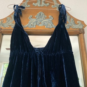 May include: A dark blue velvet dress with thin straps that tie at the shoulders. The dress has a gathered waist and a loose, flowing skirt.