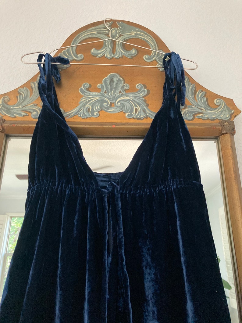 May include: A dark blue velvet dress with thin straps that tie at the shoulders. The dress has a gathered waist and a loose, flowing skirt.