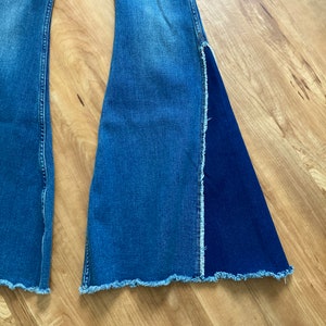 Puede incluir: Un par de pantalones vaqueros acampanados de mezclilla azul con dobladillo sin rematar. Una pierna tiene un panel de mezclilla azul oscuro contrastante.