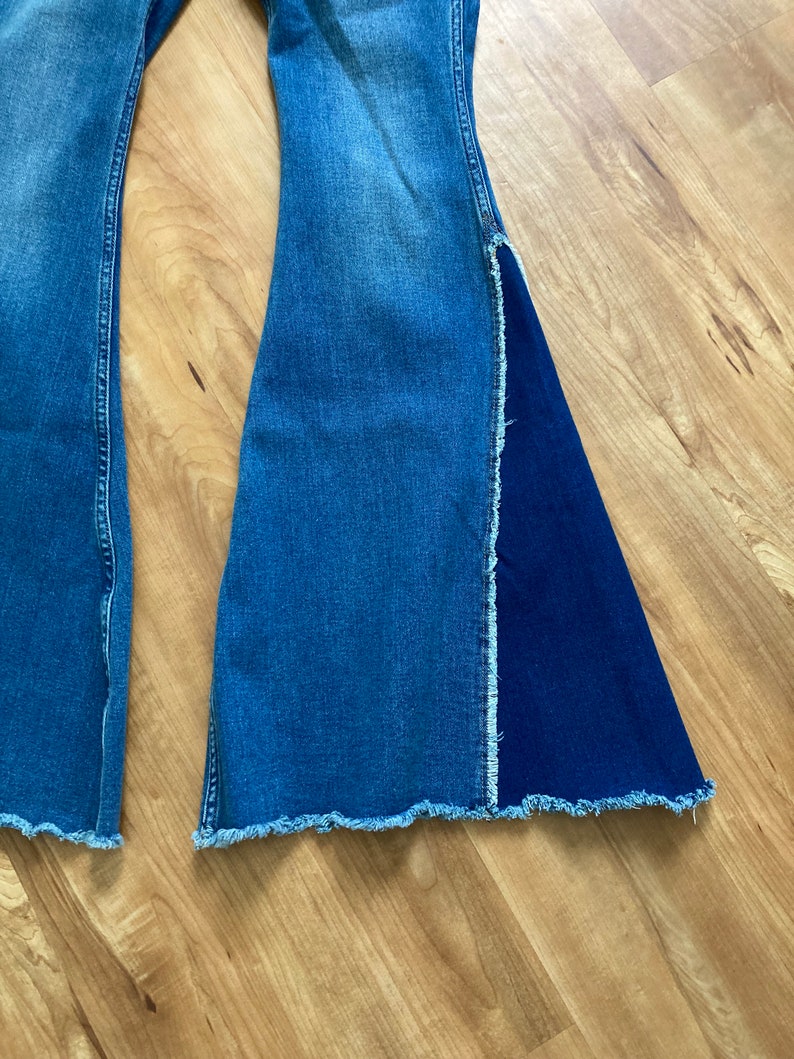 Puede incluir: Un par de pantalones vaqueros acampanados de mezclilla azul con dobladillo sin rematar. Una pierna tiene un panel de mezclilla azul oscuro contrastante.