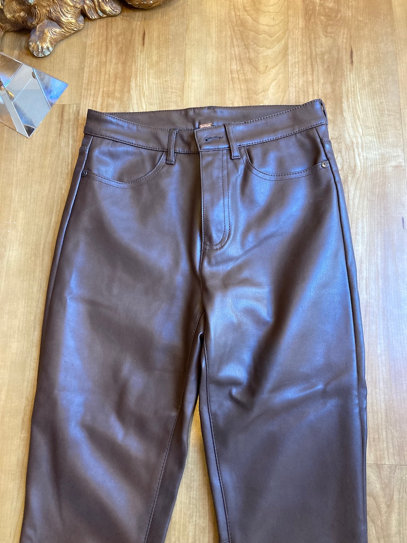 K&ouml;nnte beinhalten: Braune Kunstlederhose mit geradem Bein. Die Hose hat einen Rei&szlig;verschluss und einen Knopfverschluss.
