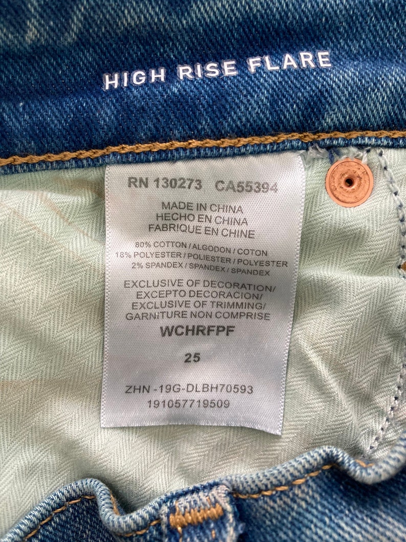 Puede incluir: Una etiqueta blanca con texto negro unida a un par de pantalones vaqueros azules. La etiqueta dice "RN 130273 CA55394 Made in China Hecho en China Fabrique en Chine 80% Cotton/Algodon/Coton 18% Polyester/Poliester/Polyester 2% Spandex/Spandex/Spandex Exclusive of Decoration/Excepto Decoracion/Exclusive of Trimming/Garniture Non Comprise WCHRFPF 25 ZHN-19G-DLBH70593 191057719509"