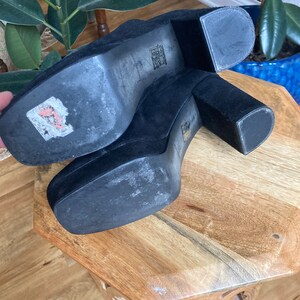May include: A pair of black suede platform heels with a thick block heel. The heels are on a wooden surface.