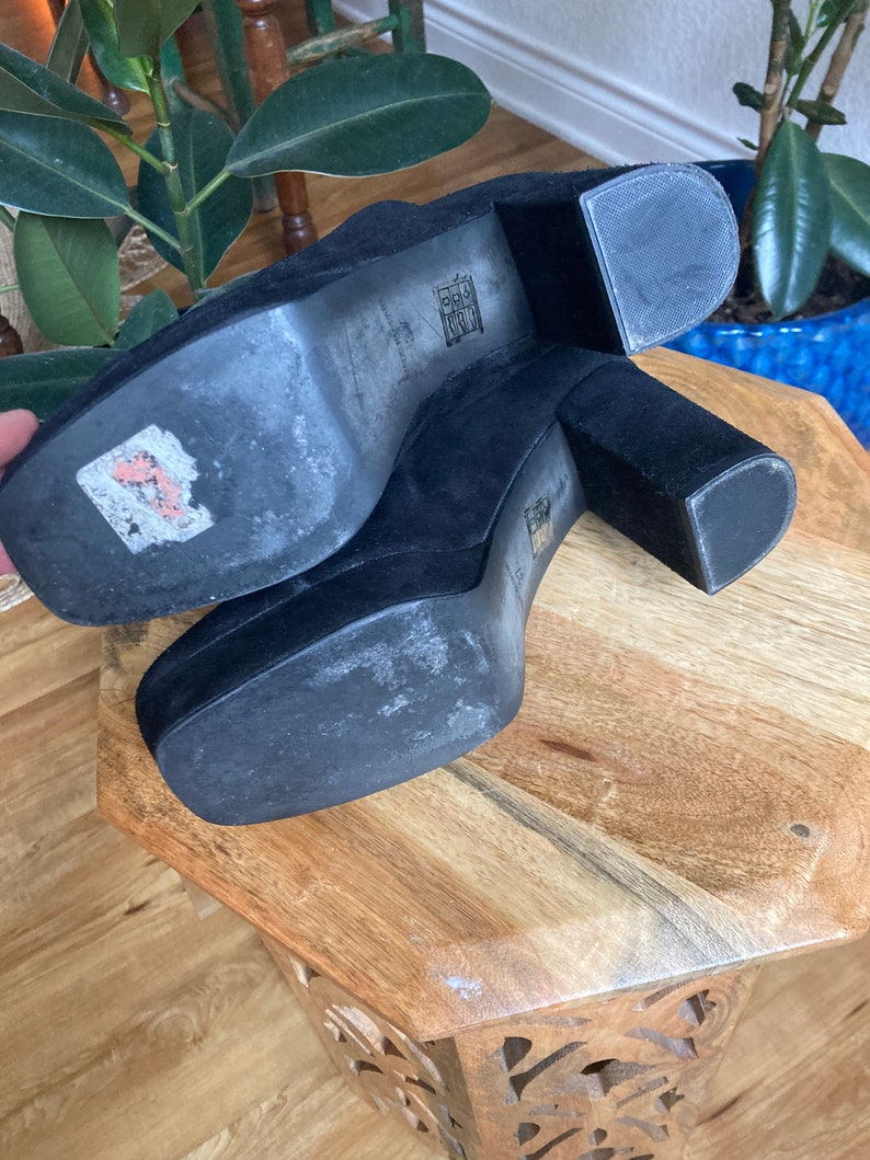 May include: A pair of black suede platform heels with a thick block heel. The heels are on a wooden surface.