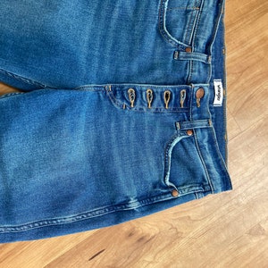 Puede incluir: Un par de pantalones vaqueros azules de mezclilla con cierre de bot&oacute;n y una etiqueta de Wrangler. Los pantalones vaqueros tienen cinco bolsillos y un lavado claro.