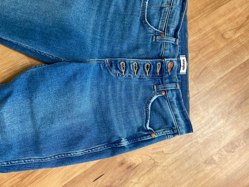Puede incluir: Un par de pantalones vaqueros azules de mezclilla con cierre de bot&oacute;n y una etiqueta de Wrangler. Los pantalones vaqueros tienen cinco bolsillos y un lavado claro.