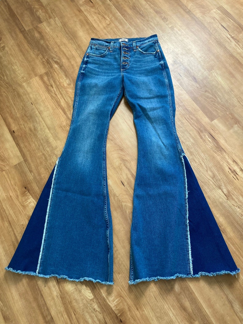 Puede incluir: Un par de pantalones vaqueros de campana azul denim con un panel de tela azul oscuro contrastante en el exterior de cada pierna. Los vaqueros tienen cierre de bot&oacute;n y dobladillo deshilachado.