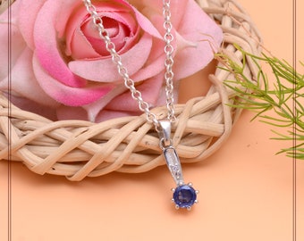 May include: A silver necklace with a pendant featuring a round, deep blue gemstone. The pendant is accented with small, clear stones and hangs from a delicate chain. The necklace is displayed on a woven basket with a pink rose in the background.