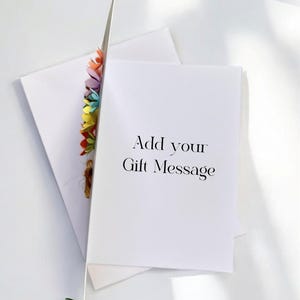 May include: A white greeting card with the text "Add your Gift Message" is propped open, revealing a colorful floral embellishment. An envelope and a sprig of green leaves are also visible.
