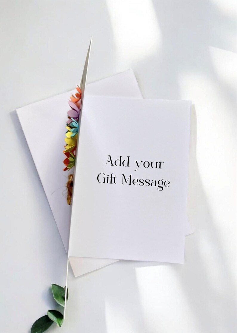 May include: A white greeting card with the text "Add your Gift Message" is propped open, revealing a colorful floral embellishment. An envelope and a sprig of green leaves are also visible.