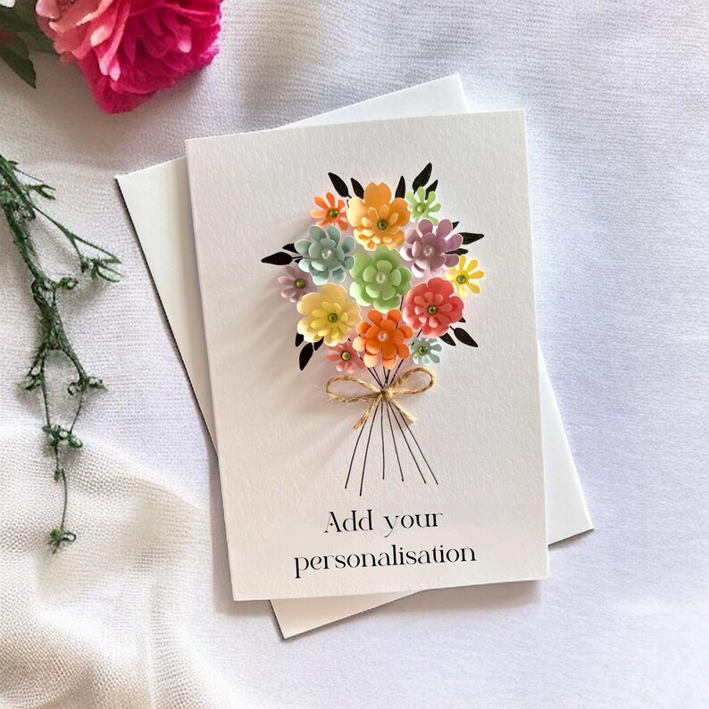 May include: A white greeting card with a colorful paper flower bouquet design. The bouquet features orange, yellow, pink, green, and blue flowers tied with twine. The text "Add your personalisation" is printed below the bouquet.