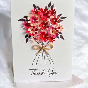 May include: A white greeting card featuring a paper flower bouquet in shades of red and pink, tied with twine. The card reads "Thank You" in a handwritten font. The card is accompanied by a white envelope.