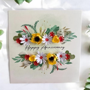 May include: A handmade anniversary card with a floral design. The card features a cream-colored background with a decorative arrangement of yellow, white, and red paper flowers, and green leaves. The words "Happy Anniversary" are written in black script.