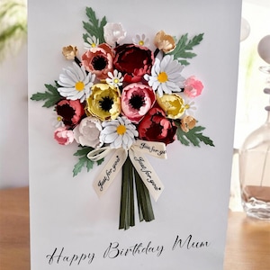 May include: A white greeting card with a paper flower bouquet. The bouquet features red, pink, yellow, and white flowers with green leaves. A ribbon with "Just for you" is tied around the stems. The card reads "Happy Birthday Mum."