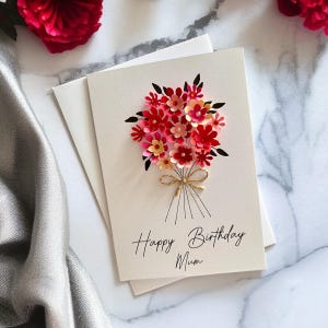 May include: A cream-colored greeting card with a paper flower bouquet in shades of red, pink, and peach. The bouquet is tied with twine and the card reads "Happy Birthday Mum" in black script. An envelope is behind the card.