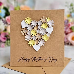 May include: A handmade Mother's Day card with a heart-shaped floral design. The heart is composed of yellow, white, and tan paper flowers, with white paper hearts. The card is on a brown background with the text "Happy Mother's Day".