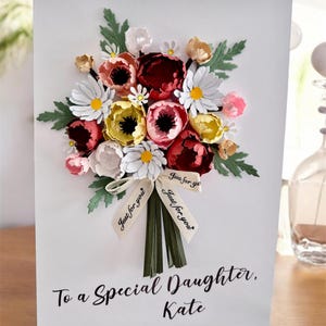 May include: A white greeting card with a paper flower bouquet. The bouquet features red, pink, yellow, and white flowers with green leaves. A ribbon with "Just for you" is tied around the stems. The card reads "To a Special Daughter, Kate."