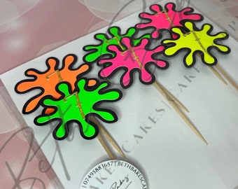 May include: Set of four neon green, pink, and yellow paint splatter cupcake toppers with gold glitter accents. The toppers are attached to wooden skewers.